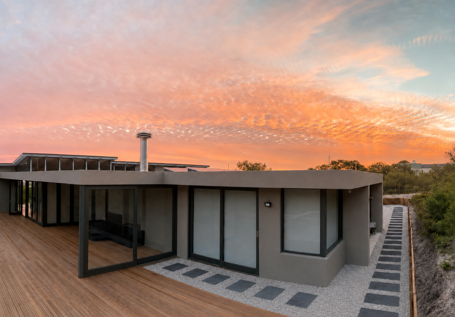 Contemporary single-storey home with timber deck, sliding glass walls and a stone pathway, beneath a vivid sunset sky